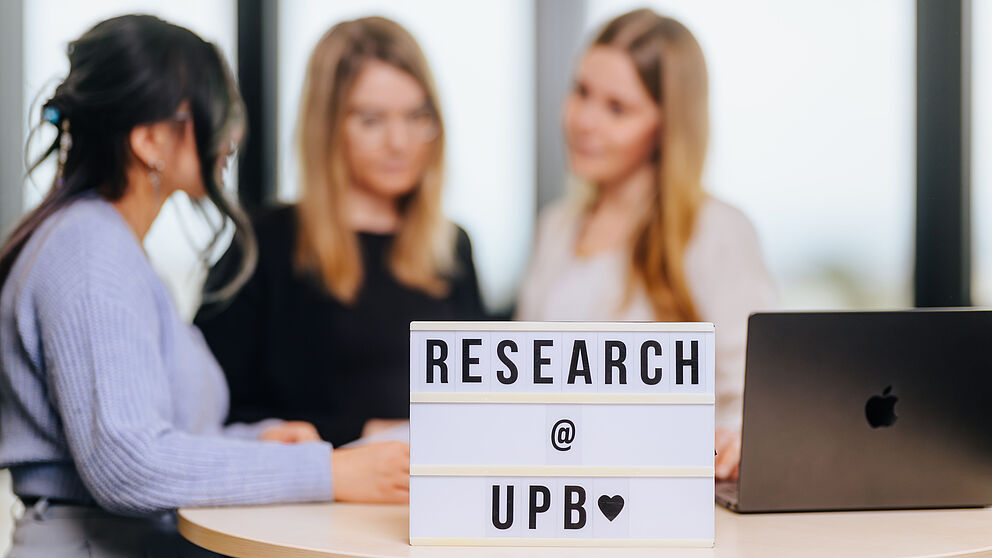 Drei Personen, im Vordergrund ein Leuchtkasten mit dem Text "Research UPB".