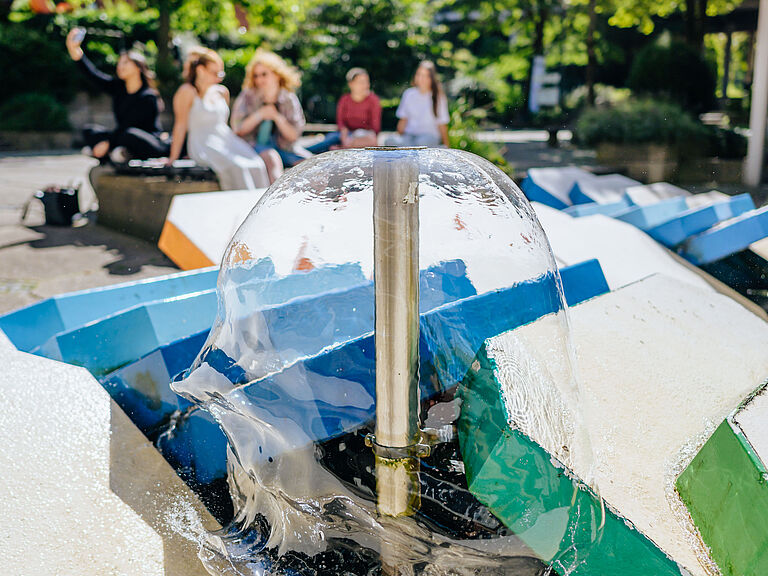 Junge Menschen sitzen zusammen und reden, im Vordergrund ein Springbrunnen.