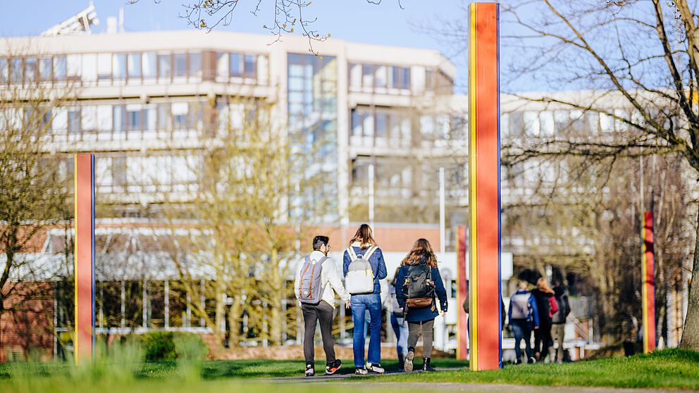 Außengelände der Universität mit einer Wiese und Menschen im Vordergrund und einem Gebäude im Hintergrund.