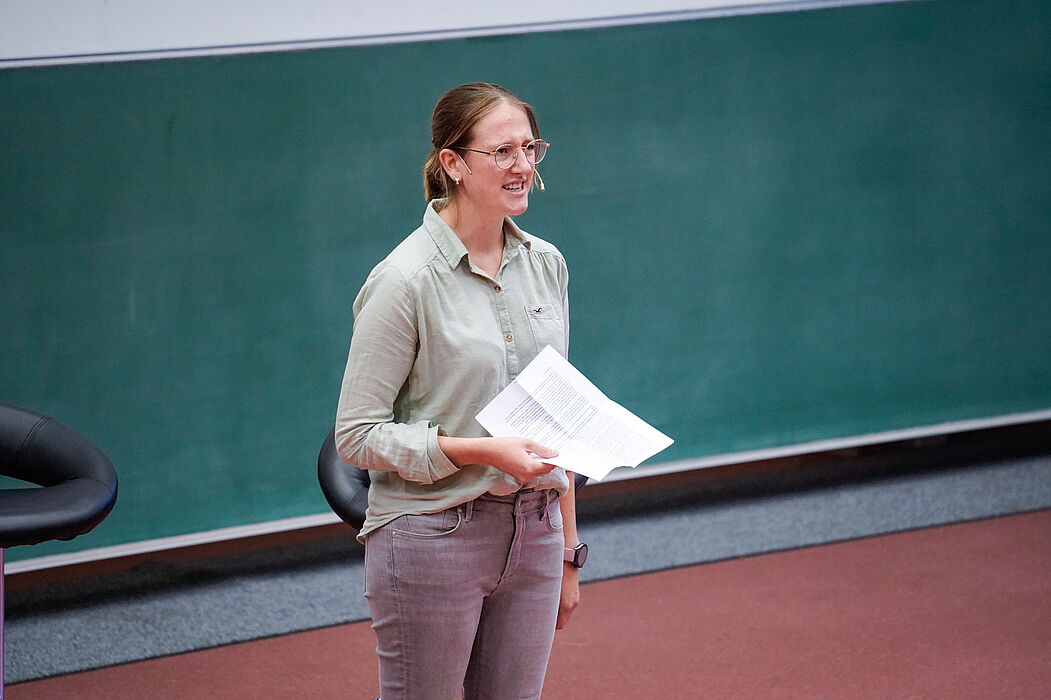 Eine junge Frau mit einem Blatt Papier in der Hand spricht in ein Headset-Mikrofon.