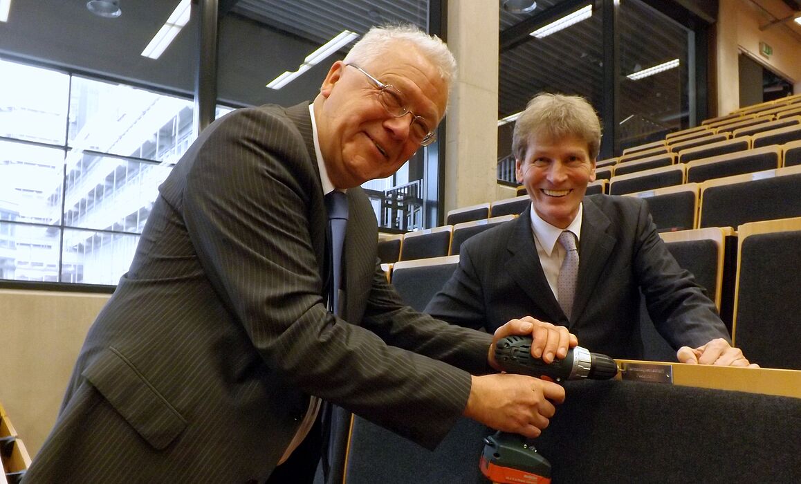 Foto (Universität Paderborn, Jasmin Rostam): Dr. Rolf Dietz von der Harmoniegesellschaft (li.) bringt die Plakette am gestifteten „Leerstuhl“ an. Prof. Dr. Nikolaus Risch freut sich über die Spende.