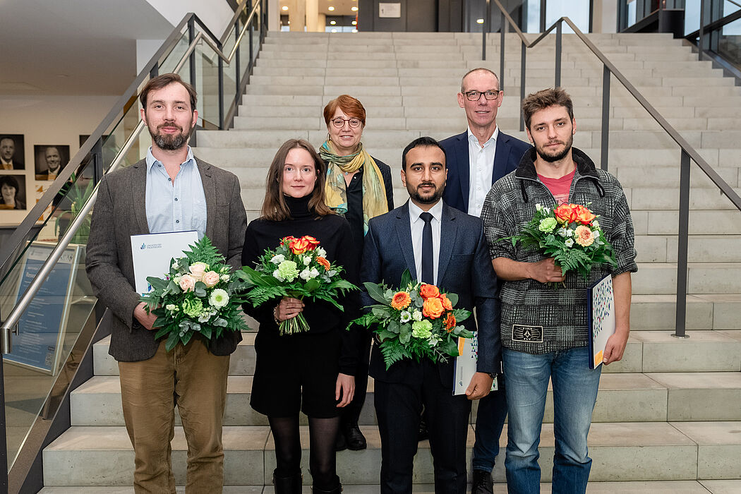 Zwei Frauen und vier Männer stehen in zwei Reihen auf einer Truppe, vier von Ihnen halten Blumensträuße.