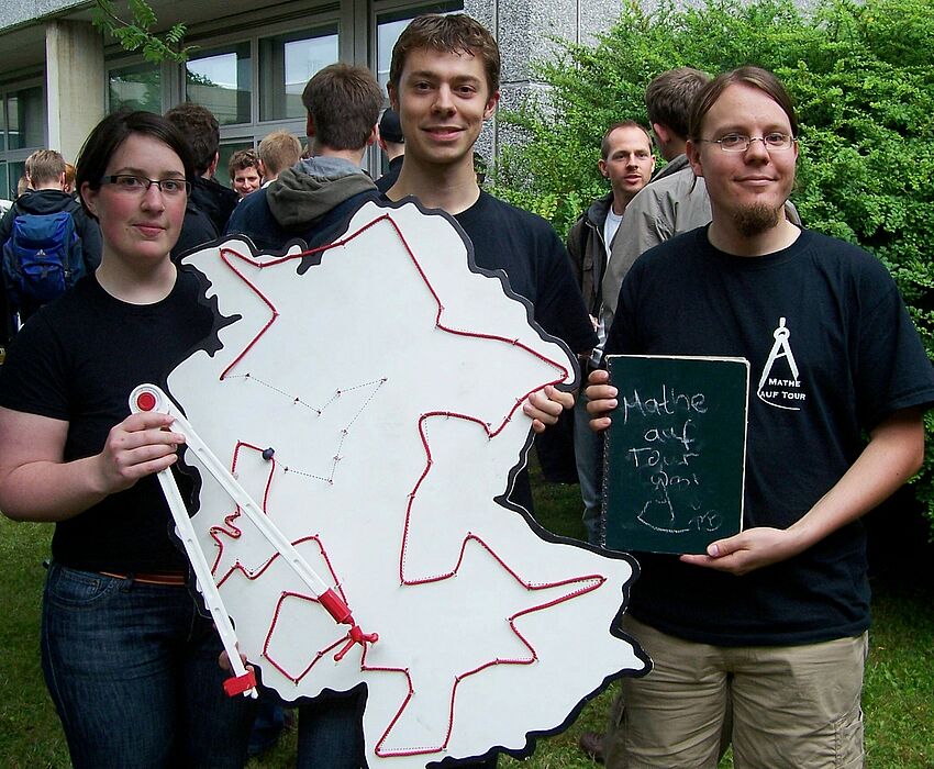 Foto (Mark Heinemann): Anna Flötotto (li.) und Jörg Zender (r.) überreichten Andreas Cord-Landwehr den Zirkel samt Reisetagebuch und Routenplan.