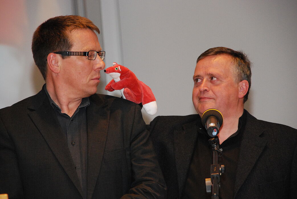 Foto (Universität Paderborn, Christoph Streicher): Hans Peter Brandl-Bredenbeck (links) assistierte Florian Söll, nebst Handpuppe, bei seiner Zugabe nach dem Sieg.