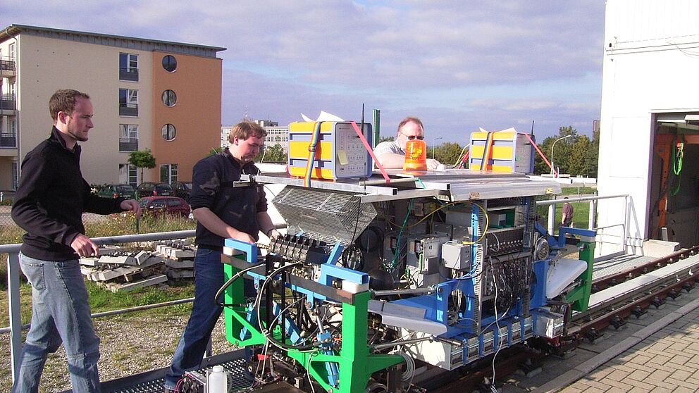 Foto (Sascha Lutters): Letzte Änderungen vor der Testfahrt: Studenten und Mitarbeiter (v. li.: Christian Kunzmann, Markus Jürgens, Hans-Joachim Kraus) des Fachgebiets Rechnernetze installieren zwei kooperativ funkende Prototypen (orange) auf dem RailCab