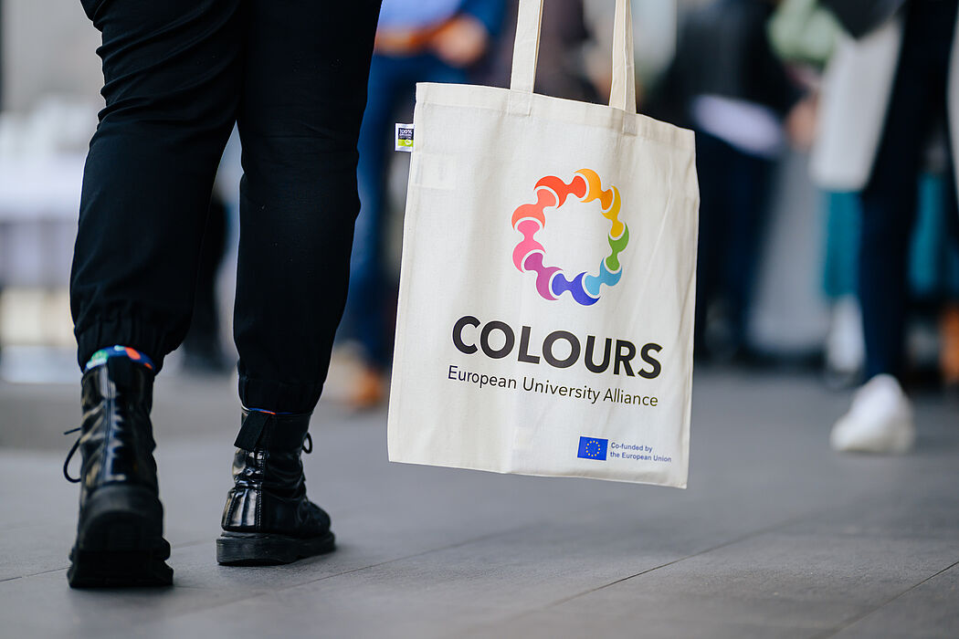 Beine in schwarzer Hose und mit schwarzen Stiefeln, daneben eine Tasche mit Aufdruck des Logos der europäischen Hochschulallianz COLOURS. 