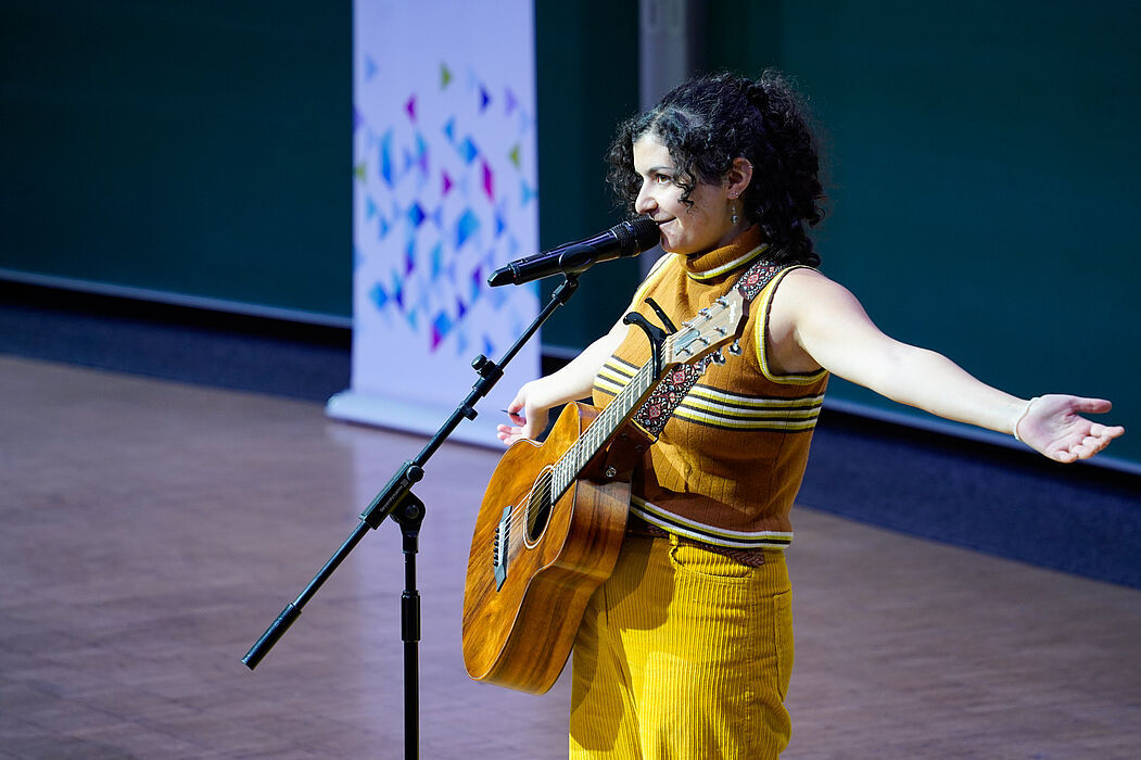 Eine junge Frau singt am Mikrofon und hat eine Gitarre umgeschnallt. 