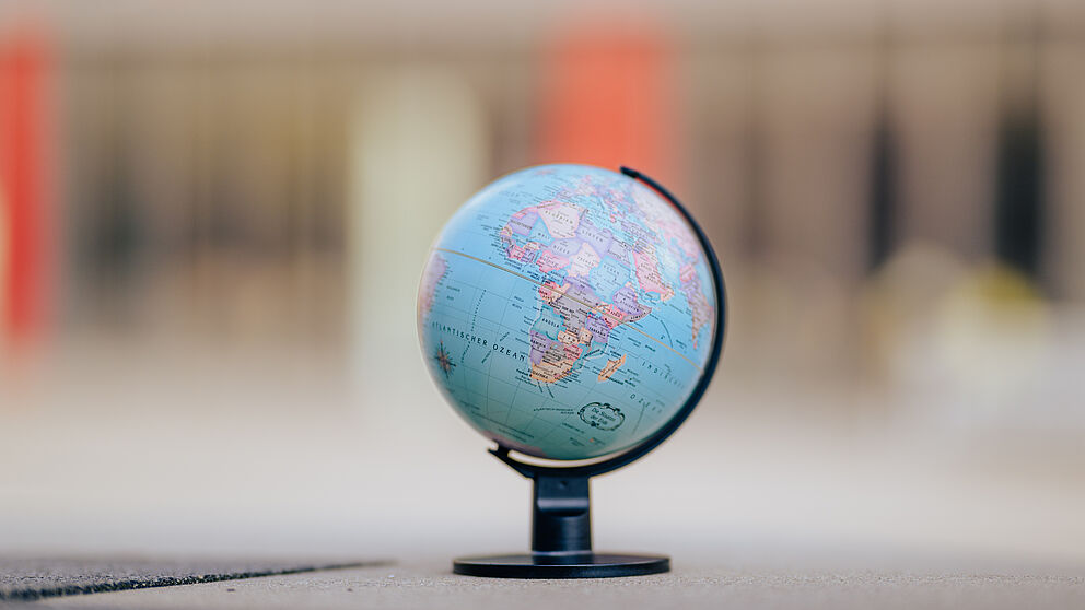 Ein Globus steht auf grauem Untergrund. Der Hintergrund ist verschwommen. Ein Gebäude ist grob zu erkennen. 