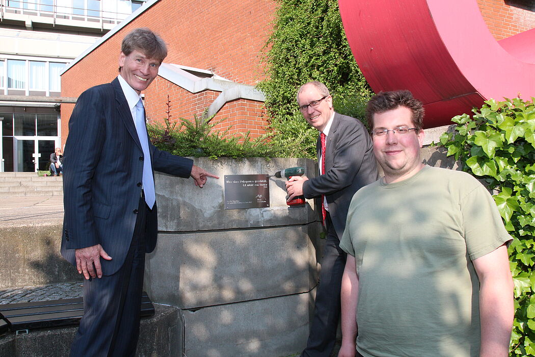Foto (Universität Paderborn, Vanessa Dreibrodt): Uni-Präsident Prof. Dr. Nikolaus Risch und Landrat Manfred Müller montieren ein Schild. Über die neuen Sitzbänke freut sich AStA-Vorsitzender Florian Rittmeier (v. l.).